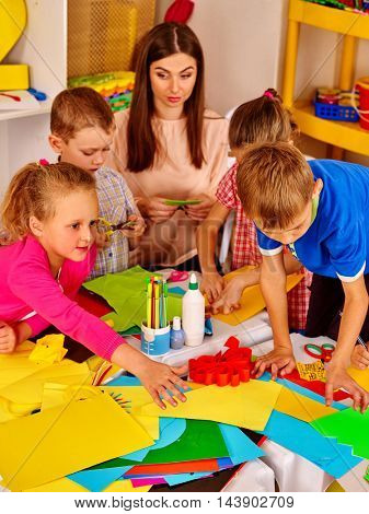Large group of children with teacher friendly making something out of colored paper on table in primary school. Children craft lesson in primary school. Childrens crafts out of paper.