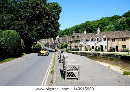 CASTLE COMBE, UNITED KINGDOM - JULY 20, 2016 - Stone cottages alongside the river Bybrook with wooden benches and a sports car in the foreground Castle Combe Wiltshire England UK Western Europe. July 20, 2016.