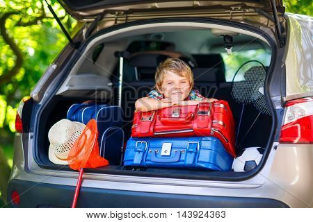 Adorable little kid boy sitting in car trunk just before leaving for summer vacation with his parents. Happy child with suitcases and toys going on journey.