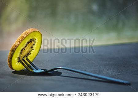 A Thin Transparent Slice Of Fresh Kiwi On A Small Black Shiny Dessert Fork On A Matte Painted Dark S