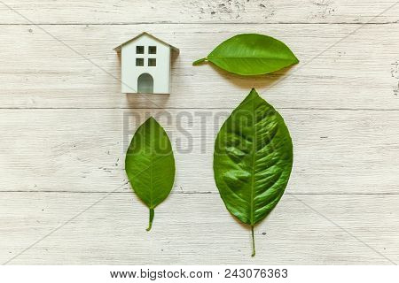 Miniature White Toy Model House With Green Leaves On Wooden Backgdrop. Eco Village, Abstract Environ