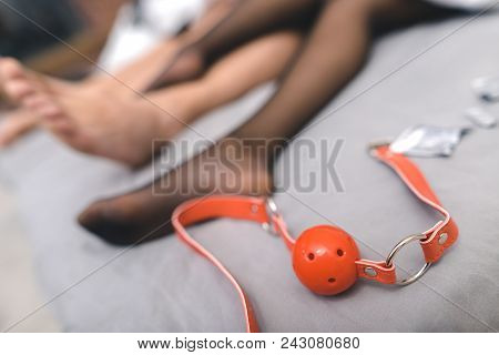 Close Up Of Red Ball-gag Near Legs On Bed. Couple In Love Making Love In Bedroom. Sex Party Concept.