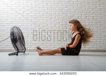 Little Girl Struggling With Heat And Sitting In Front Of A Fan With Flying Hair In An Apartment On A