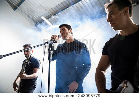 Male Lead Singer With Two Guitar Players Performing Music On Stage With Smoke And Light At Back