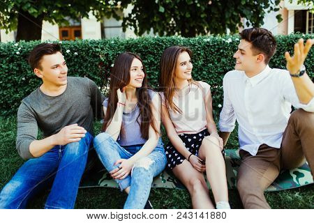 Group Of Four Young Friends In An Urban Park Sitting On The Grass In The Summer Sunshine Laughing An
