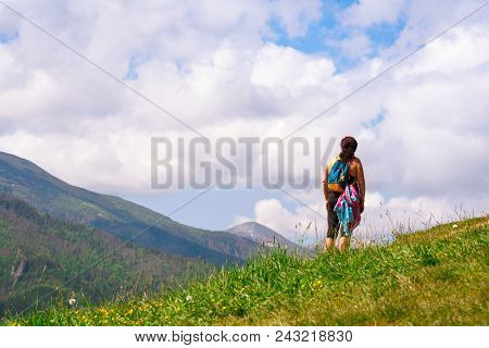 An Adult Woman With A Backpack Stands With Her Back To The Camera On The Green Grass In The Mountain