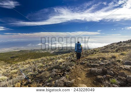 Man With Backpack On Path In Semidesert Near Neide Volcano On Tenerife Island With Stones And Blue S