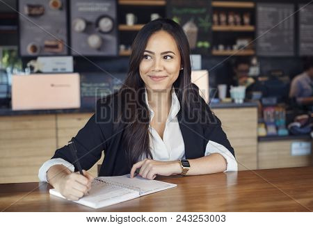 Beautiful Hispanic Young Woman In Suit Working In The Cafe Writing In A Copybook O Diary, Looking At