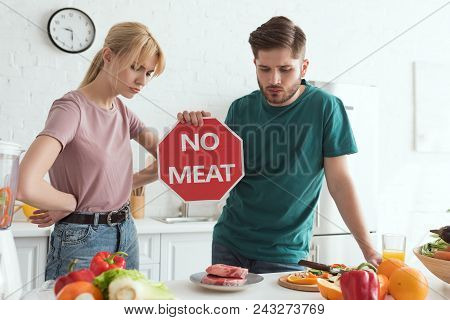 Vegan Couple With No Meat Sign At Table With Raw Meat And Vegetables In Kitchen At Home