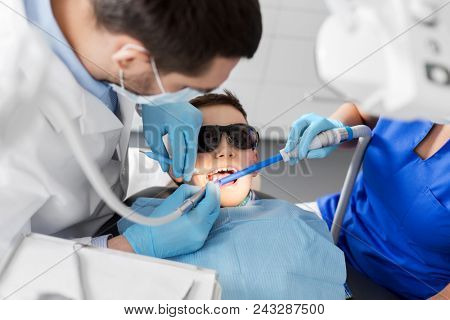 medicine, dentistry and healthcare concept - dentist and assistant with dental drill and saliva ejector treating kid patient teeth at dental clinic