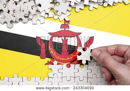 Brunei Darussalam Flag  Is Depicted On A Table On Which The Human Hand Folds A Puzzle Of White Color