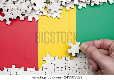 Guinea Flag  Is Depicted On A Table On Which The Human Hand Folds A Puzzle Of White Color.