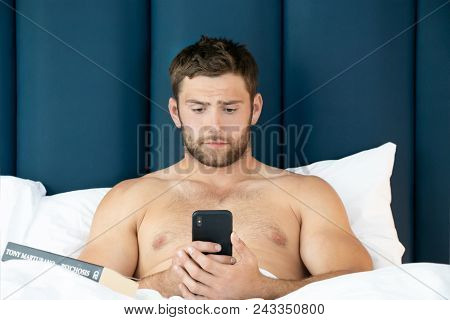 Handsome Man With Abs, Muscular, Hunky Body And Beard Lies Naked In Between White Sheets On Bed Usin