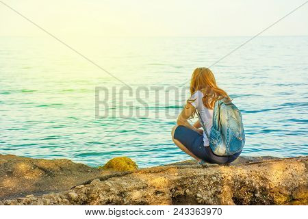 A Girl With Red Hair And A Backpack Is Squatting On A Stone By The Sea And Looking Out Into The Dist