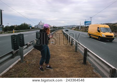 Pretty Young Woman Hitchhiking Along A Road. Girl With Rose Hat Hitchhikng During Tourist Season.
