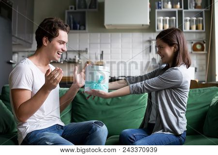 Smiling Girl Giving Gift Box To Excited Boyfriend Making Present For His Birthday Sitting On Sofa At