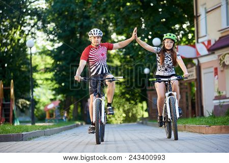Cyclist Couple In Cycling Clothing And Helmets Giving High Fives Riding Bikes Down Paved Street In P