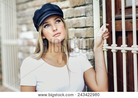 Young Blonde Woman Wearing Cap Standing Near A Brick Wall.