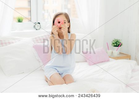 Small Pretty Female Child Holds Tasty Sweet Doughnut, Poses In Bedroom On Comfortabled Bed, Dressed 