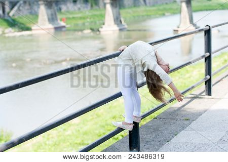 Kid Girl Bends Backwards As Flexible Gymnast Near Railing At Riverside Background. Girl Kid Gymnast 