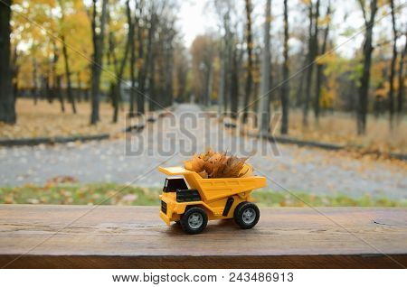 A Small Toy Yellow Truck Is Loaded With Yellow Fallen Leaves. The Car Stands On A Wooden Surface Aga