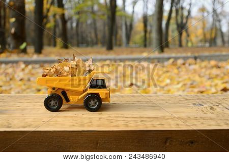 A Small Toy Yellow Truck Is Loaded With Yellow Fallen Leaves. The Car Stands On A Wooden Surface Aga