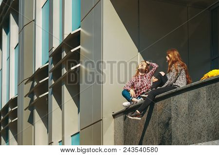 College Students Take A Break After Classes. Young Girls With Reddish Hair Sit On A Modern Building 