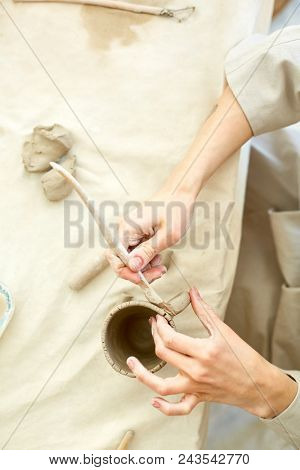 Overview of craftswoman hands with handtool for making earthenware finishing up with clay mug
