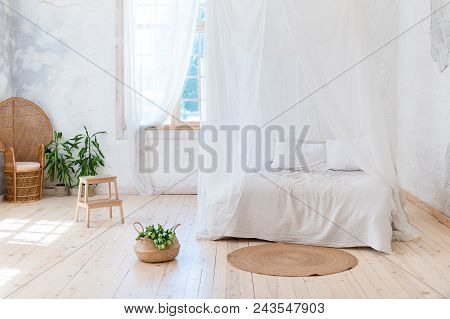 Cozy Bedroom In Light Colors With A Wooden Floor, A Large Four-poster Bed, Wicker Chair And Basket O