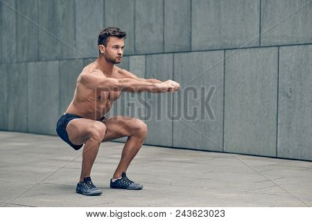 Fit Muscular Shirtless Young Man Working Out In Town Doing Squats In A Forecourt Of A Commercial Bui
