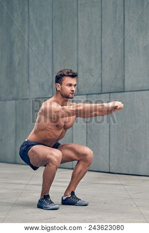 Fit Muscular Shirtless Young Man Working Out In Town Doing Squats In A Forecourt Of A Commercial Bui
