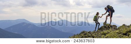 Young Tourists With Backpacks, Athletic Boy Helps Slim Girl To Clime Rocky Mountain Top Against Brig