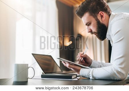 Young Bearded Businessman In Shirt Sitting At Desk In Front Of Laptop, Making Notes In Digital Table