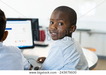 African young boy using computer in class. Portrait of happy schoolboy sitting at desk with laptop. Smiling black child looking at camera while classmate working on computer desktop. 