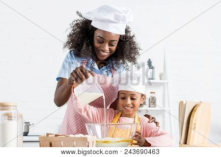 African American Mother And Daughter In Chef Hats Pouring Milk In Bowl With Ingredientes For Dough