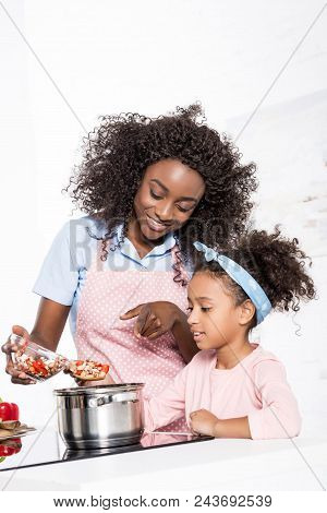 Smiling African American Mother And Daughter Putting Ingredientes In Saucepan On Kitchen