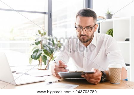 Portrait of concentrated smart businesslike man 30s in white shirt holding and looking at clipboard with paper documents during work in office