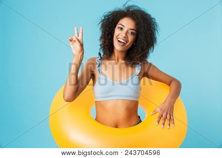 Portrait of an excited african girl dressed in swimsuit holding infatable ring and showing peace gesture isolated over blue background
