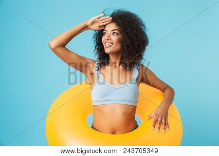 Portrait of a young african girl dressed in swimsuit holding infatable ring and looking far away isolated over blue background