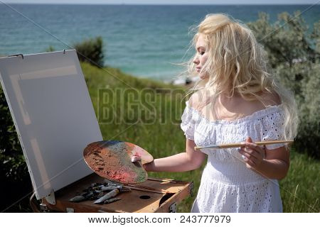 Beautiful Girl Draws A Picture In The Park Using A Palette With Paints And A Spatula. Easel And Canv