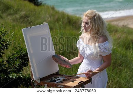 Beautiful Girl Draws A Picture In The Park Using A Palette With Paints And A Spatula. Easel And Canv