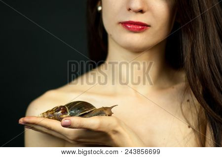 Naked Woman With Red Sensual Lips Holding On Hand A Big Snail Akhatin On Black Background In Studio.