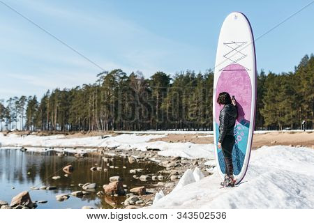 Slender Woman In Black Wetsuit Staying On The Coast And Holds Sup Board Vertically. Preparation For 