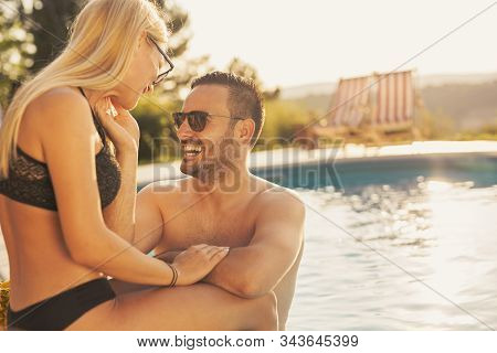 Couple In Love At A Poolside Summer Party, Sitting At The Edge Of A Swimming Pool, Sunbathing, Drink