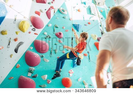Father And Teenager Son At Indoor Climbing Wall Hall. Boy Is Hanging On The Rope Using A Climbing Ha