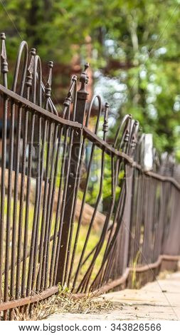 Vertical Close Up Of Old And Rusty Iron Fence Against Grassy Slope Ground And Lush Trees