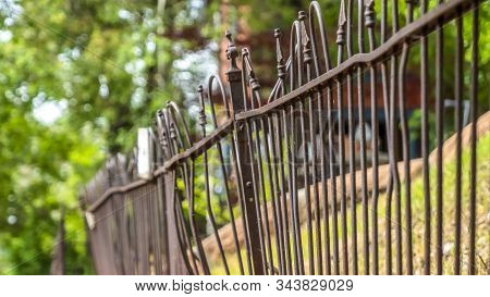 Pano Frame Close Up Of Old And Rusty Iron Fence Against Grassy Slope Ground And Lush Trees