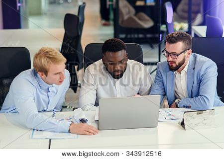 Three Businessmen Sitting At The Modern Office Tables And Analysing Data From Report And Computer To