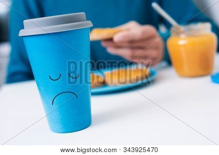 closeup of a blue cup of coffee with a sad face drawn in it on a white table and a caucasian man, wearing a blue sweater, in the background having the breakfast