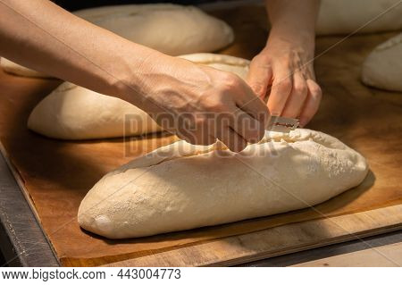 Close-up Of A Bakers Female Hands Cutting A Loaf Of Dough Into A Loaf Of Bread With A Blade Before B